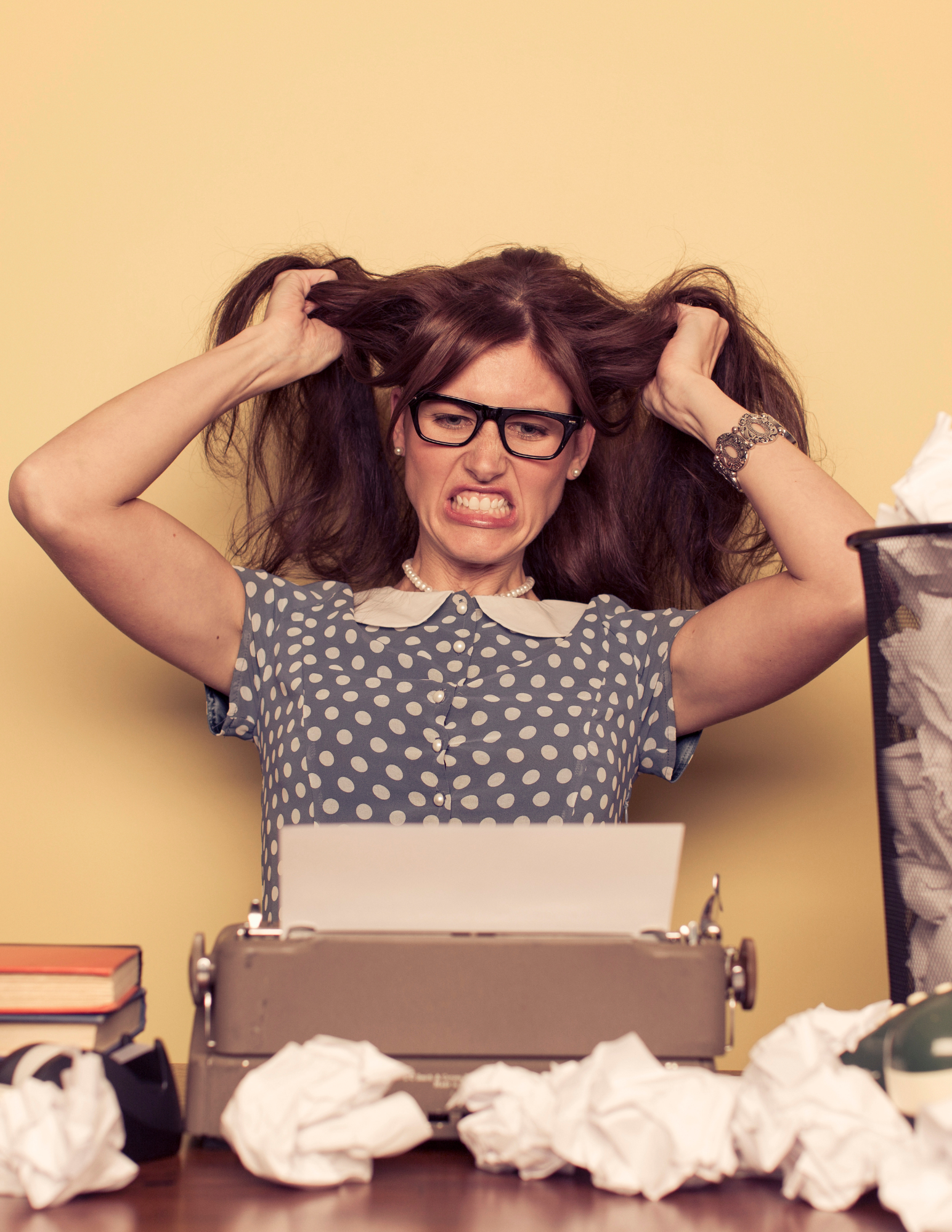 A woman wearing glasses looks frustrated and pulls her hair while sitting in front of a vintage typewriter.